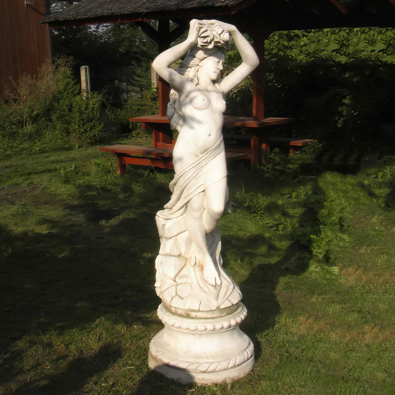 Scultura bianca in pietra di donna in piedi che tiene un cesto sulla testa in un giardino