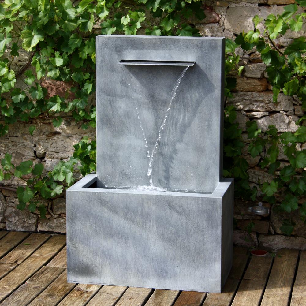 Wasserflieung aus einem modernen, grauen Gartendekobrunnen vor einer bepflanzten Wand