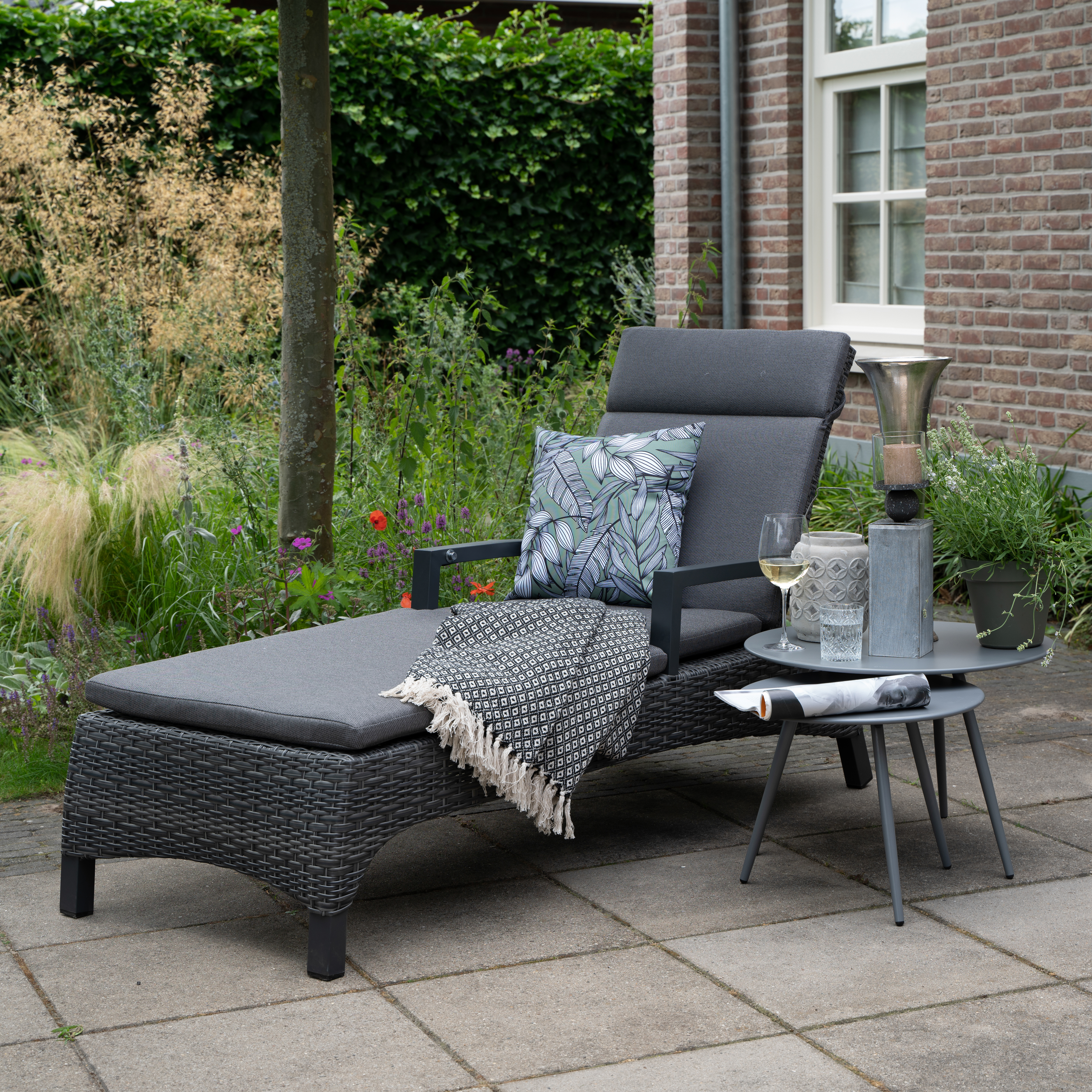 Bequeme Gartenliege mit grauen Polstern und dekorativen Kissen auf einer Terrasse vor einem Backsteinhaus