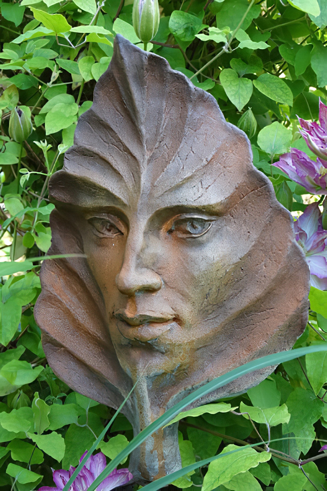 Metallgesicht in Blattform als dekorative Gartenskulptur umgeben von grünen Pflanzen und lila Blumen
