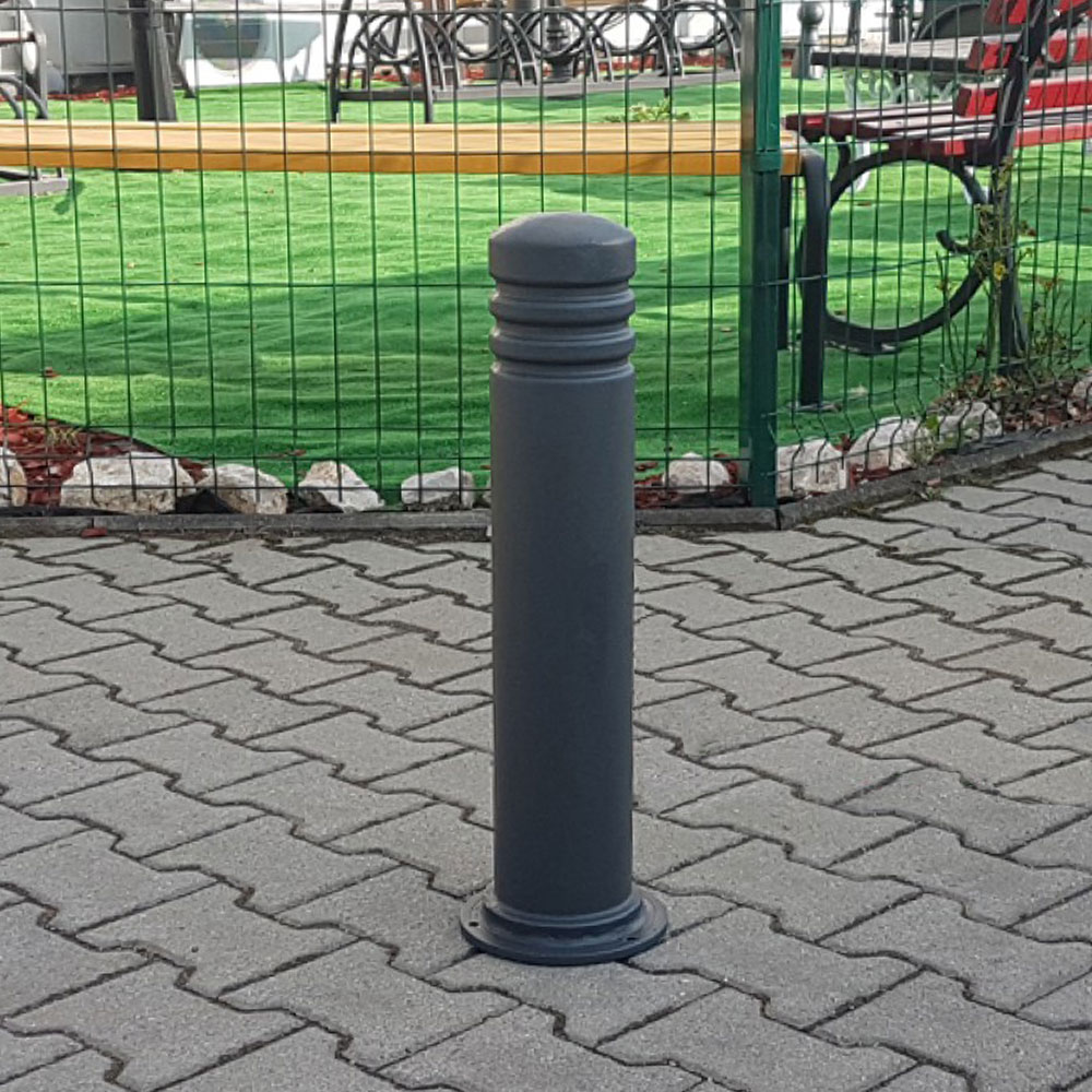 Grauer Poller aus Metall auf gepflastertem Boden vor grünem Gartenzaun