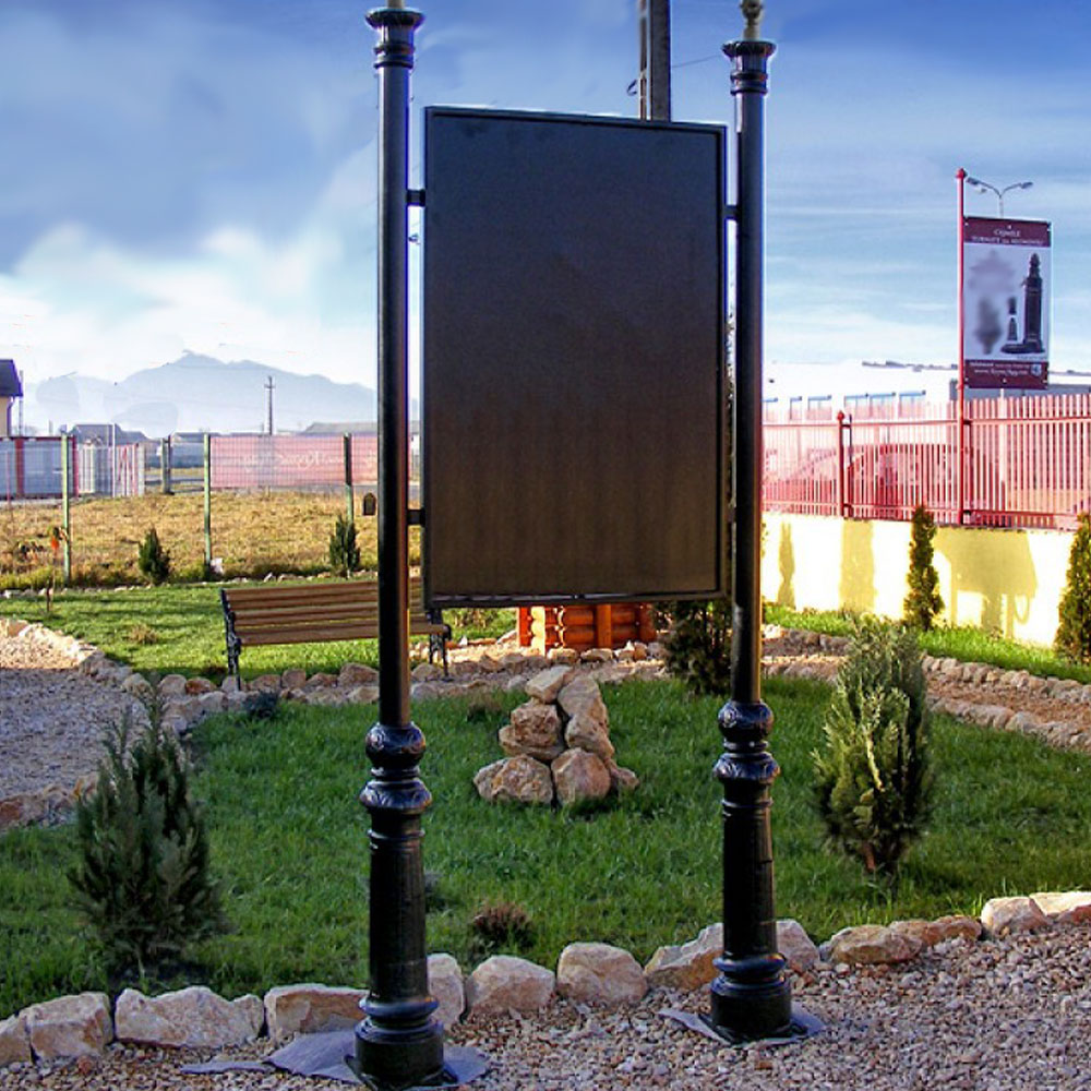 Cartello informativo nero su due colonne in un giardino con piante, sentieri e panchine