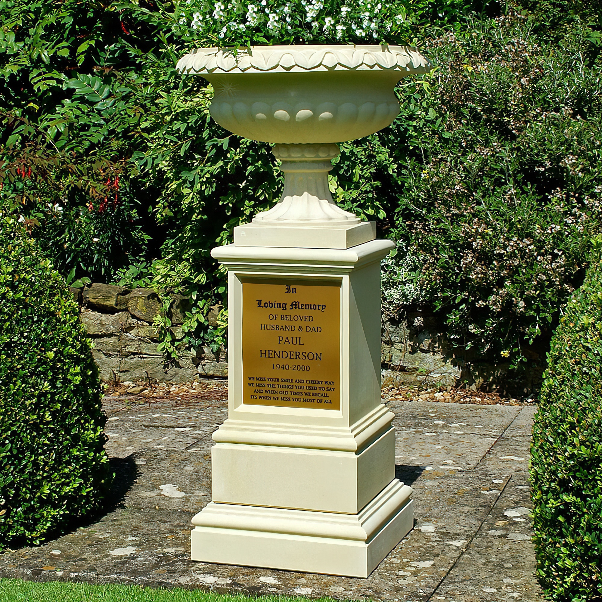 Weißer Gartenbrunnen auf Säule steht in grünem Garten mit Sträuchern
