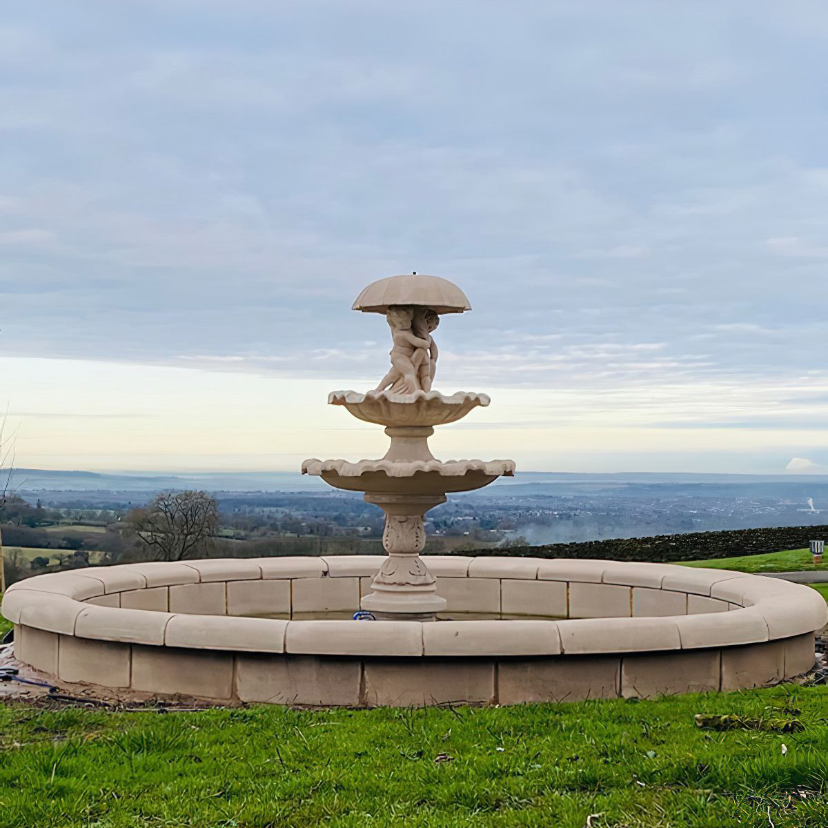 Steinerner Gartenbrunnen mit mehreren Becken und einer Statue, umgeben von grünem Gras unter bewölktem Himmel