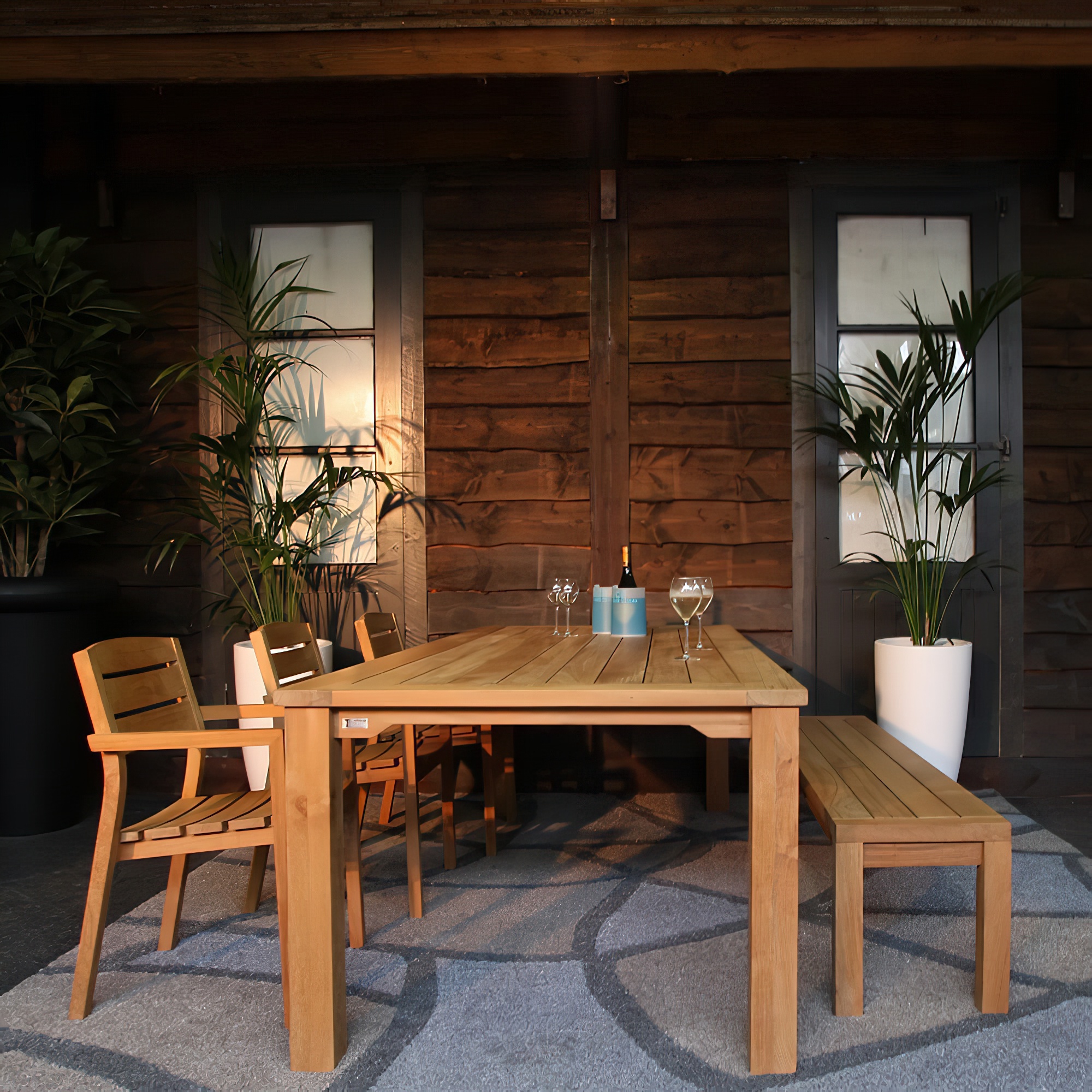 Table de jardin en bois avec chaises assorties et un banc devant un mur en bois, décorée avec des plantes