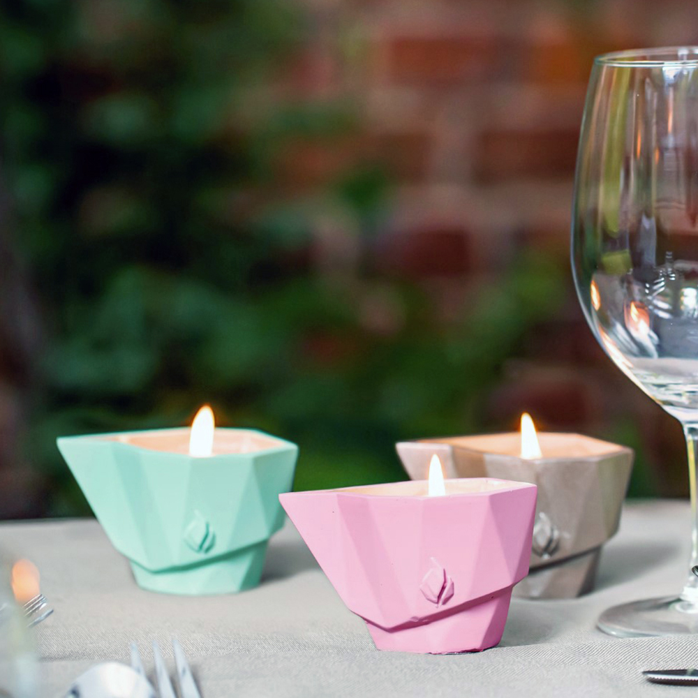 Trois bougies géométriques colorées allumées sur une table de jardin avec fond vert flou