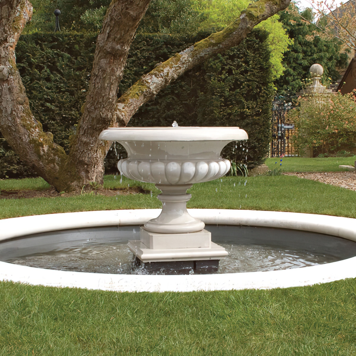 Eleganter weisser Gartenbrunnen mit fließendem Wasser umgeben von Rasen und Bäumen