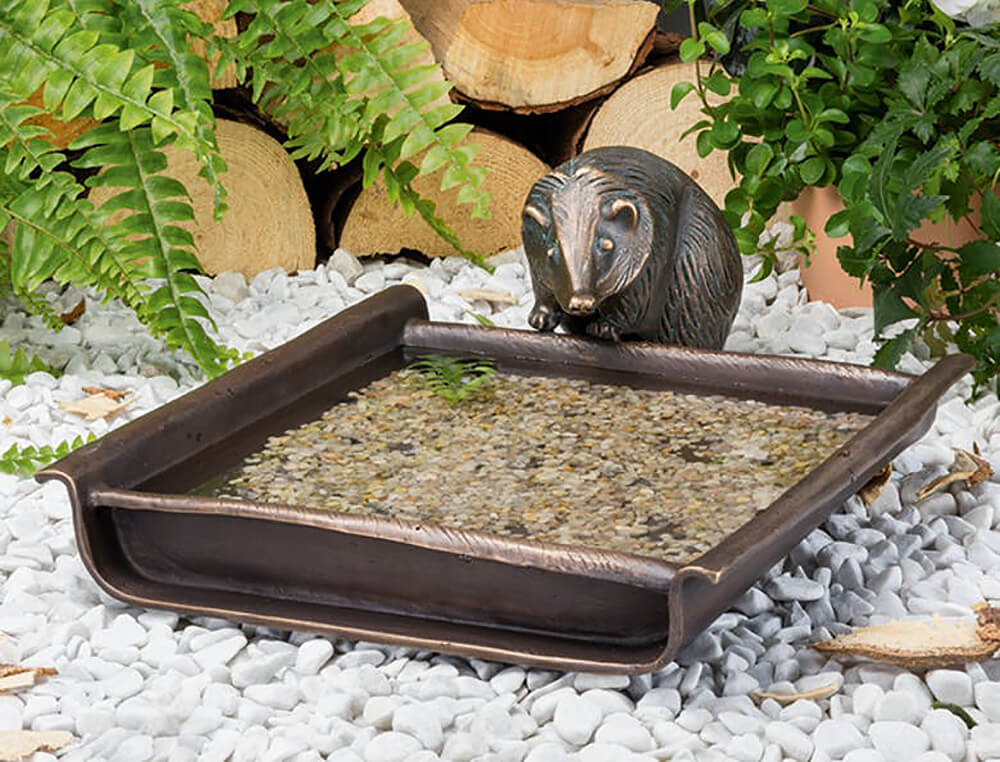 Fontana da giardino di colore bronzo con vasca d'acqua rettangolare e scultura di orso, circondata da piante verdi e elementi in legno