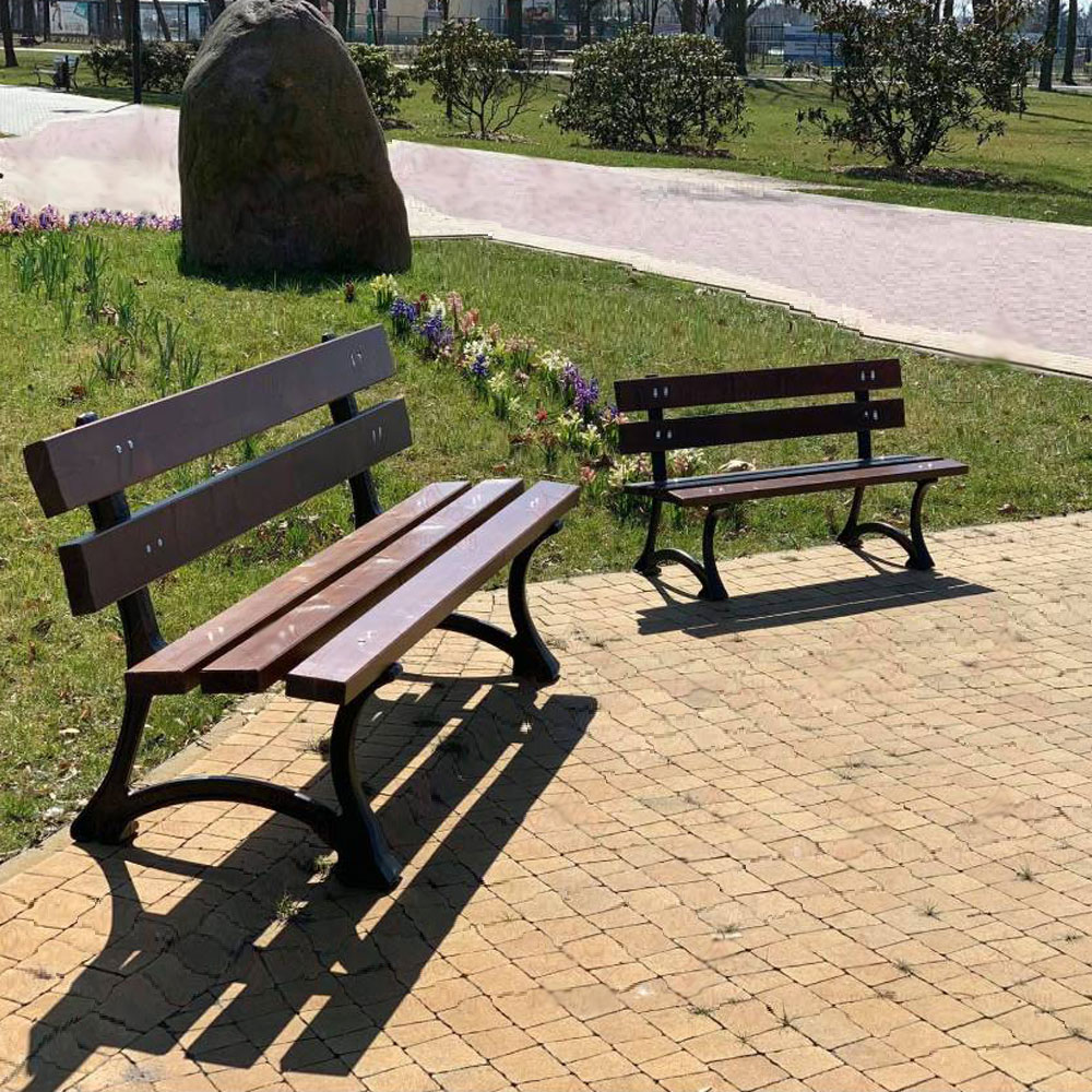 Zwei Gartenbänke aus Holz und Metall stehen auf gepflastertem Weg in einem grünen Park