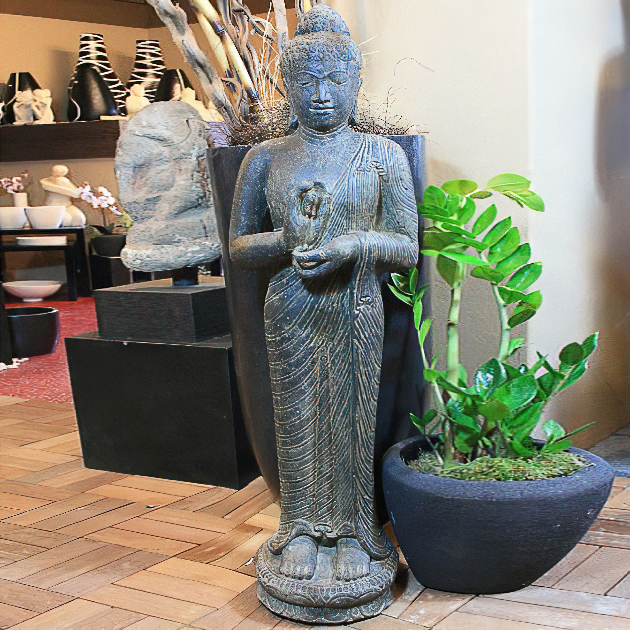 Grande statue de Bouddha debout en pierre à côté d'un pot de plante verte sur un sol en bois dans un magasin de meubles de jardin