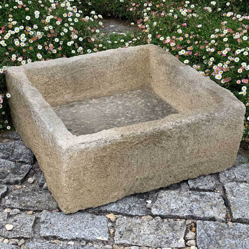Grande fontaine de jardin carrée en pierre sur des pavés avec des fleurs en arrière-plan