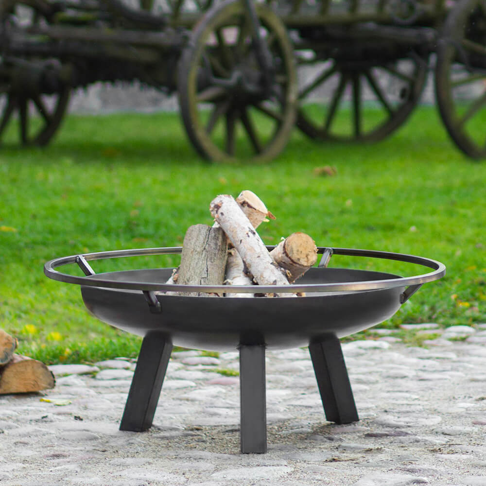 Schwarze Feuerschale mit Holzscheiten auf Steinboden im Garten mit grüner Wiese im Hintergrund