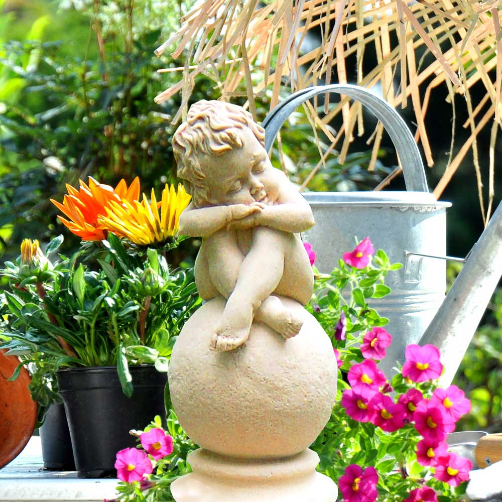 Figurine d'ange en pierre assise sur boule, entourée de fleurs colorées et de plantes
