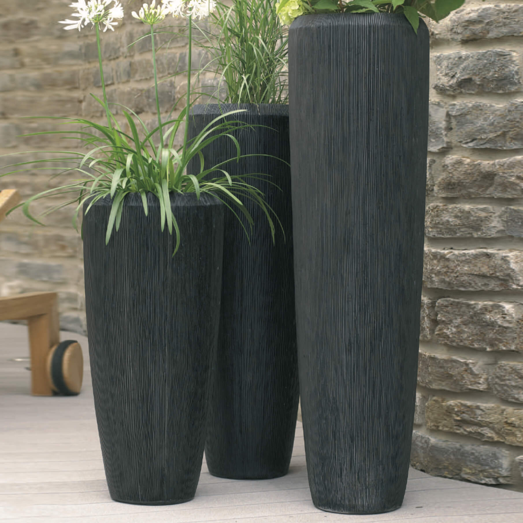 Trois grands pots de fleurs noirs avec des plantes vertes devant un mur en pierre