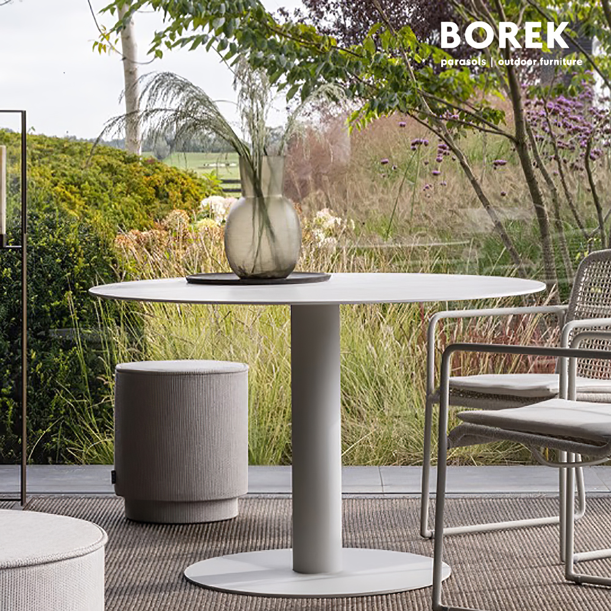 Runder weißer Gartentisch mit Vase, Hocker und Stuhl auf Terrasse mit Blick auf Garten