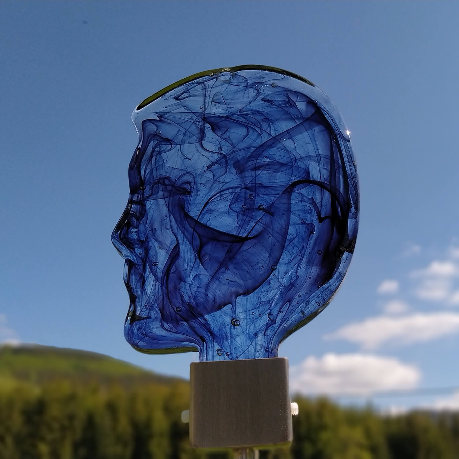 Palo da giardino in vetro blu a forma di testa con cielo azzurro e paesaggio verde sullo sfondo