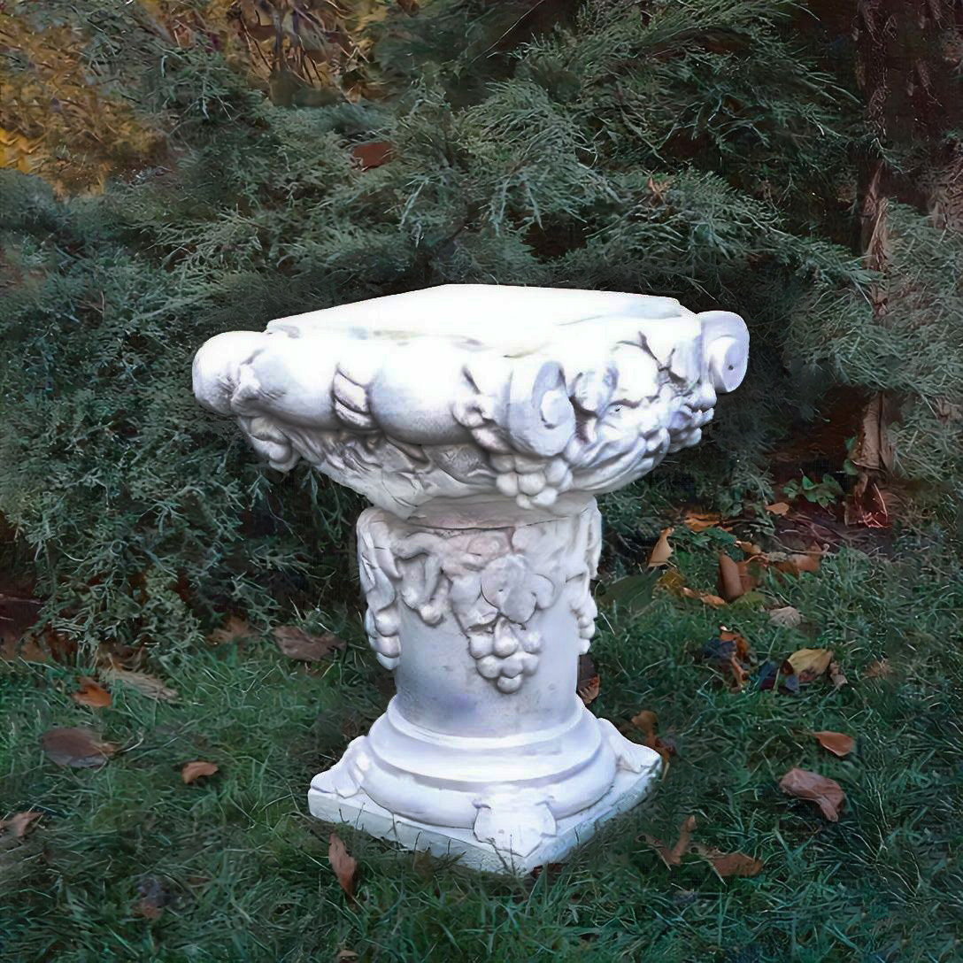 Weisse Vogeltränke aus Stein mit dekorativen Frucht- und Blattmotiven im Garten auf grünem Rasen