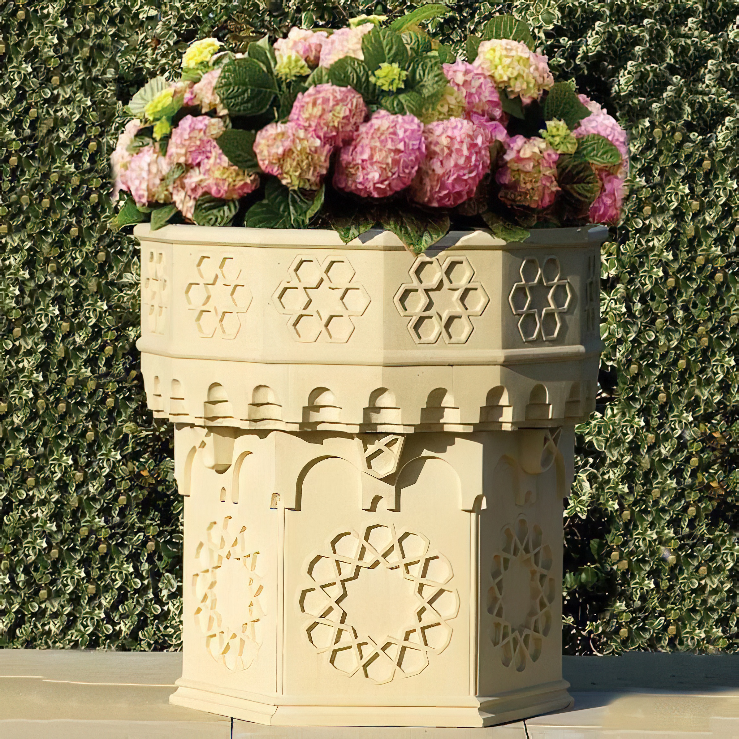 Pot de fleurs en pierre beige avec des fleurs et des décorations devant une haie