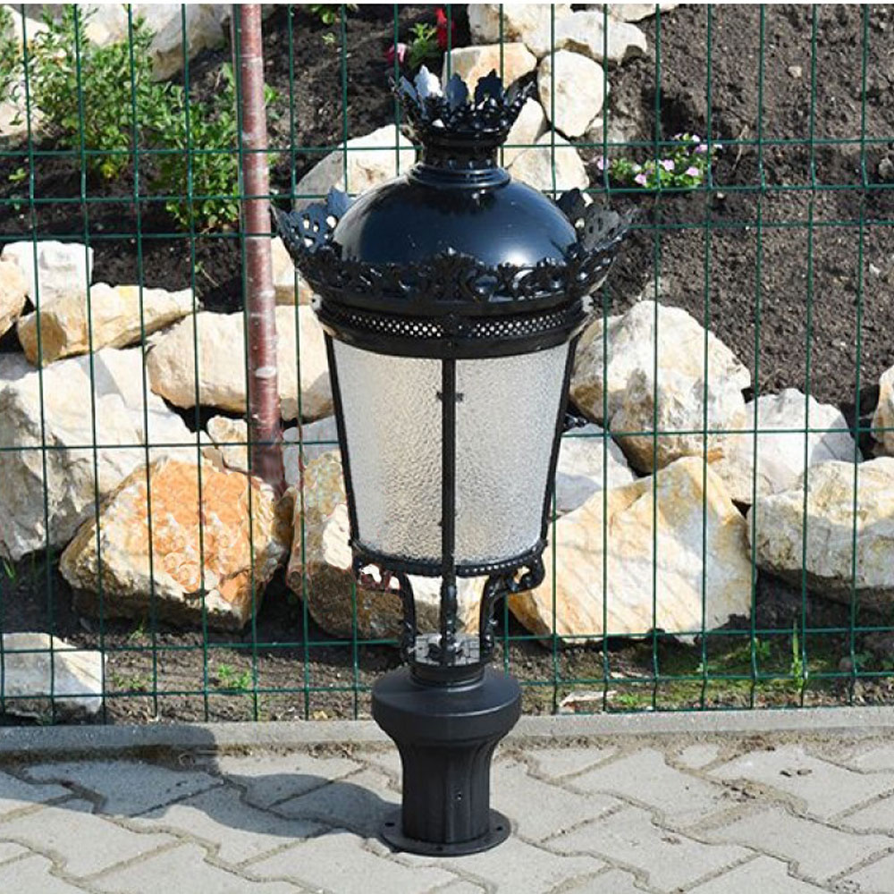 Lanterne de jardin noire en métal avec couronne décorative devant un mur de pierre et une grille en fil de fer