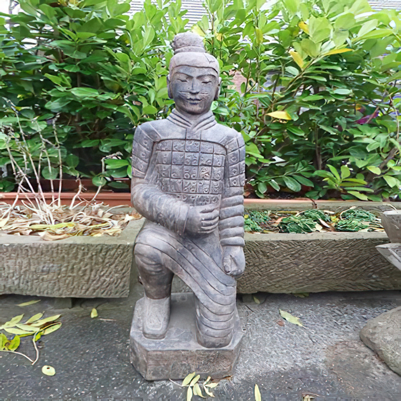 Sculpture de jardin en pierre représentant un guerrier à genoux devant des plantes vertes