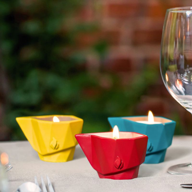 Trois bougies colorées jaune, rouge et bleue sur une table de jardin avec un verre en arrière-plan