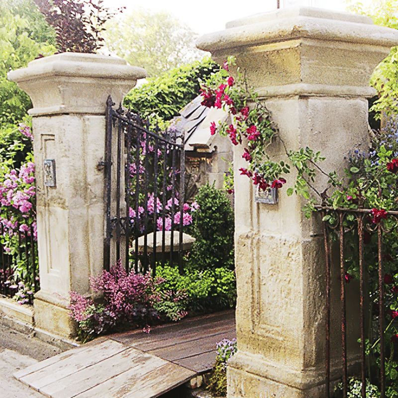 Porte de jardin en fer forgé ouverte entre des colonnes en pierre avec des plantes en fleurs