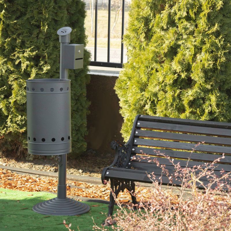 Ceneriera in metallo nero con contenitore per rifiuti accanto a una panchina da giardino sotto cespugli