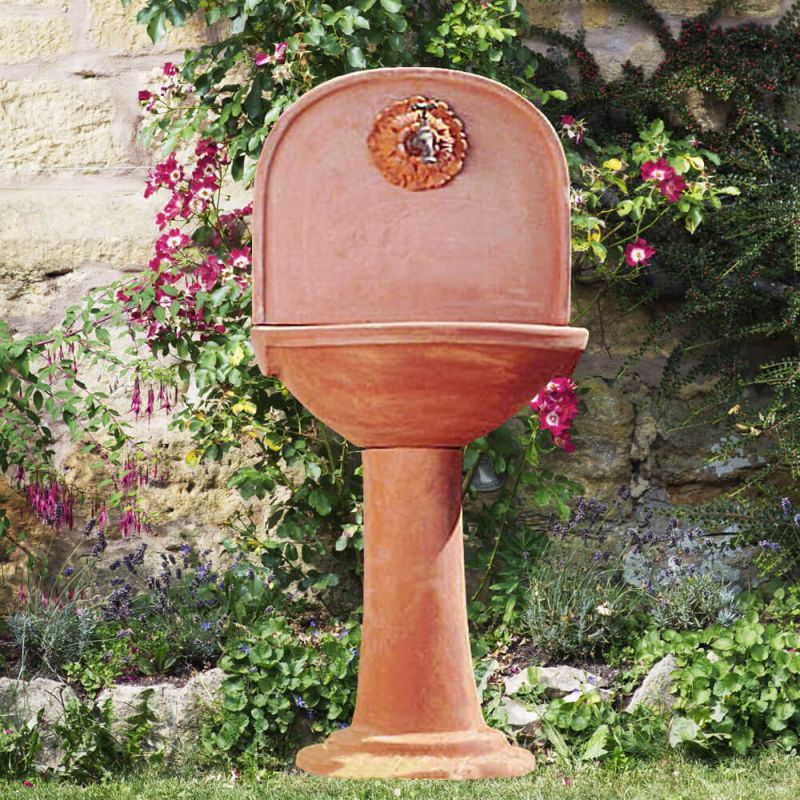 Fontana da giardino in terracotta con testa di leone e fiori davanti a un muro di pietra