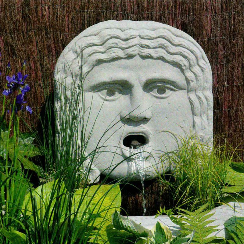 Scultura in pietra a forma di volto che funge da fontana nel giardino con piante e erba