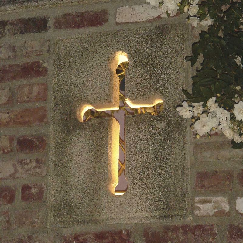 Croce di pietra retroilluminata in un muro del giardino al crepuscolo, circondata da fiori