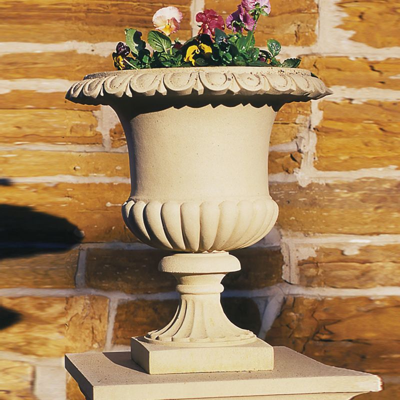 Pot de jardin en pierre beige avec des fleurs devant un mur de briques dans un jardin