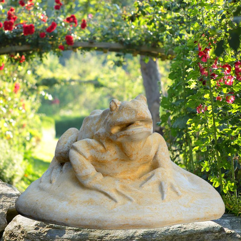 Primo piano di una scultura di rana in pietra su una pietra in un giardino verde e fiorito
