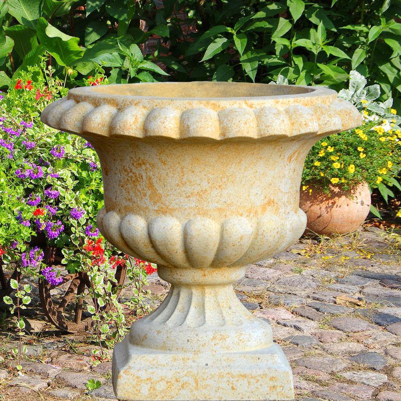 Vase en pierre beige sur un chemin pavé entouré de fleurs colorées et de feuillage vert
