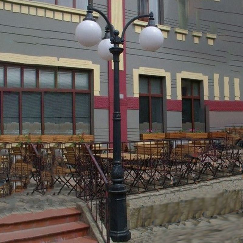 Lanterne de jardin noire avec quatre lampes blanches rondes à côté de meubles de jardin devant un bâtiment