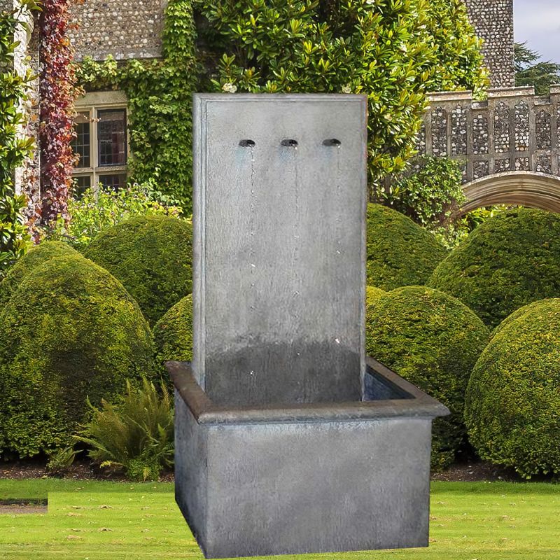Fontaine de jardin rectangulaire en métal moderne sur pelouse devant un jardin soigné