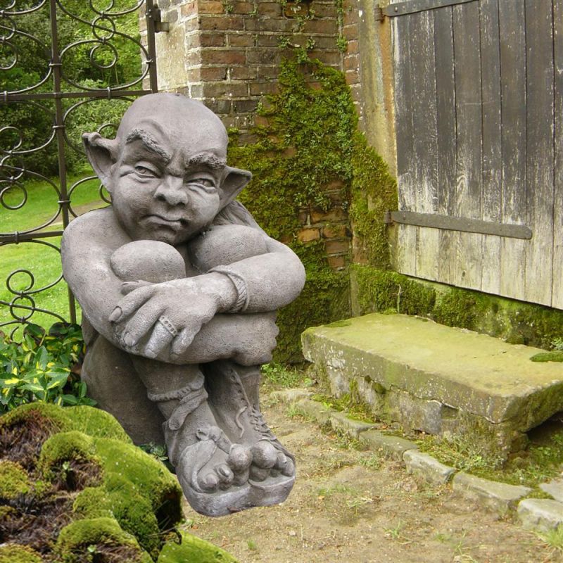 Sculpture de jardin en pierre d'un lutin assis près d'un mur en pierre et d'une porte