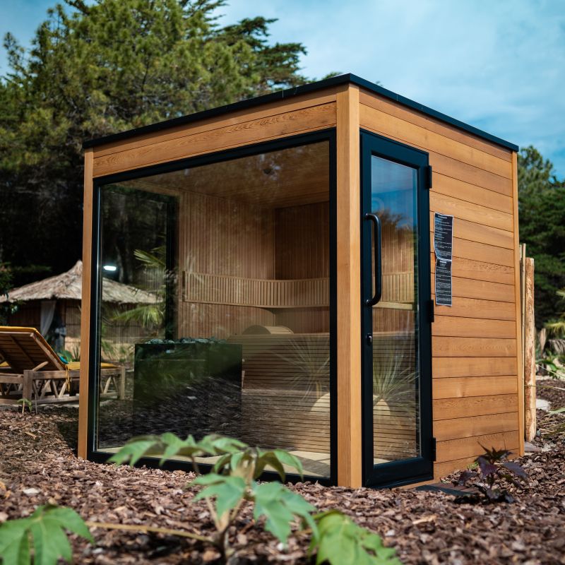 Sauna da giardino cubica in legno con grande porta vetrata e cielo azzurro sullo sfondo