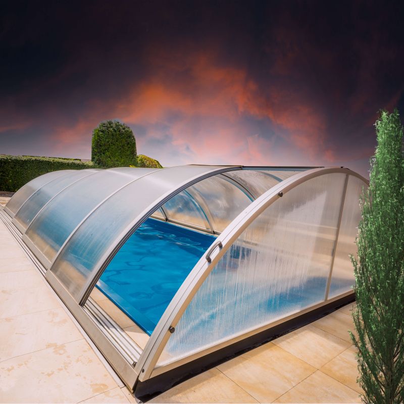 Couverture transparente de piscine protégeant la piscine de jardin, entourée de plantes de jardin sous un ciel dramatique