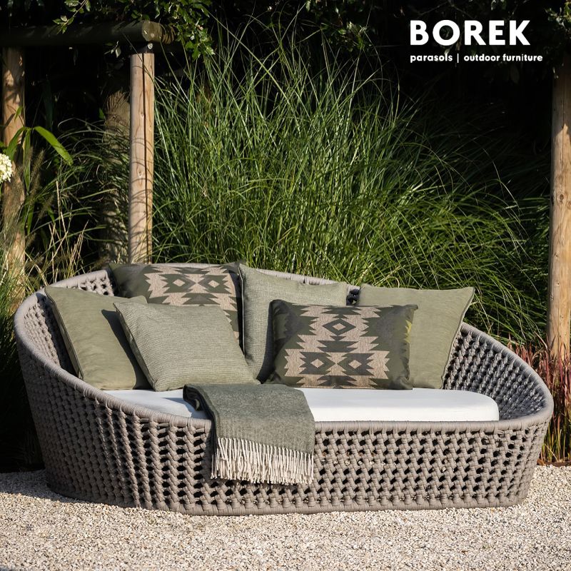 Grand fauteuil de jardin rond en rotin gris avec coussins verts et couverture devant un fond de verdure