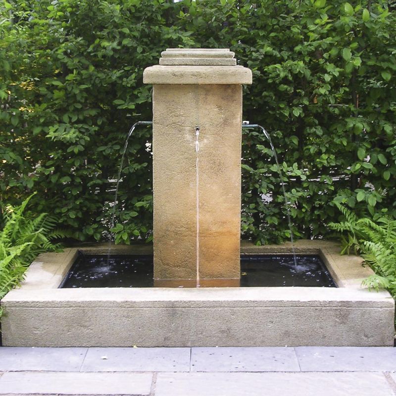 Une fontaine de jardin rectangulaire avec une colonne d'eau en pierre claire devant une haie verte