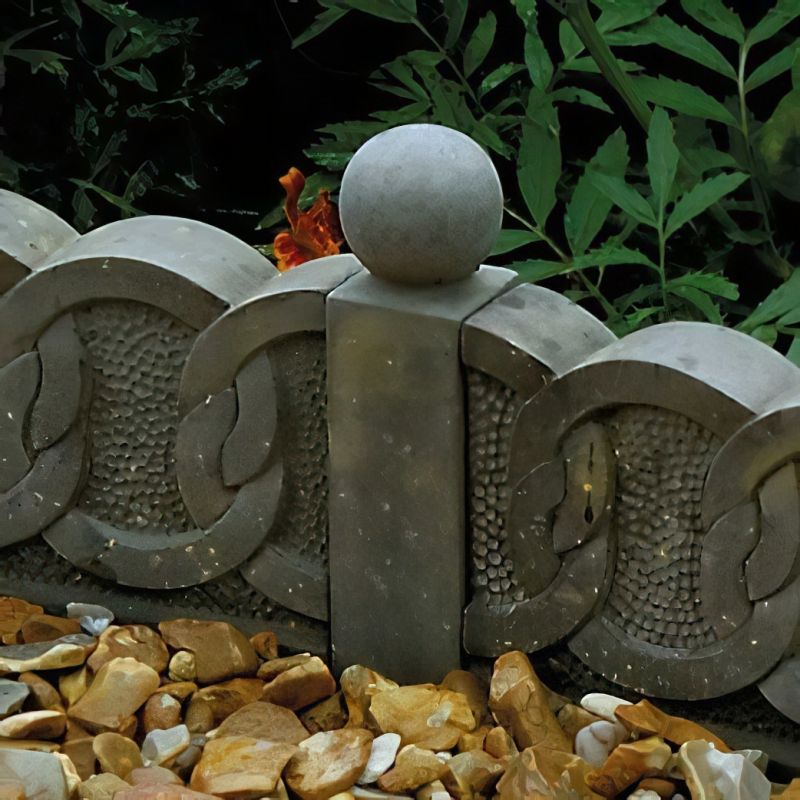 Gros plan sur un élément de clôture de jardin en pierre grise avec une boule, entouré de pierres et de plantes
