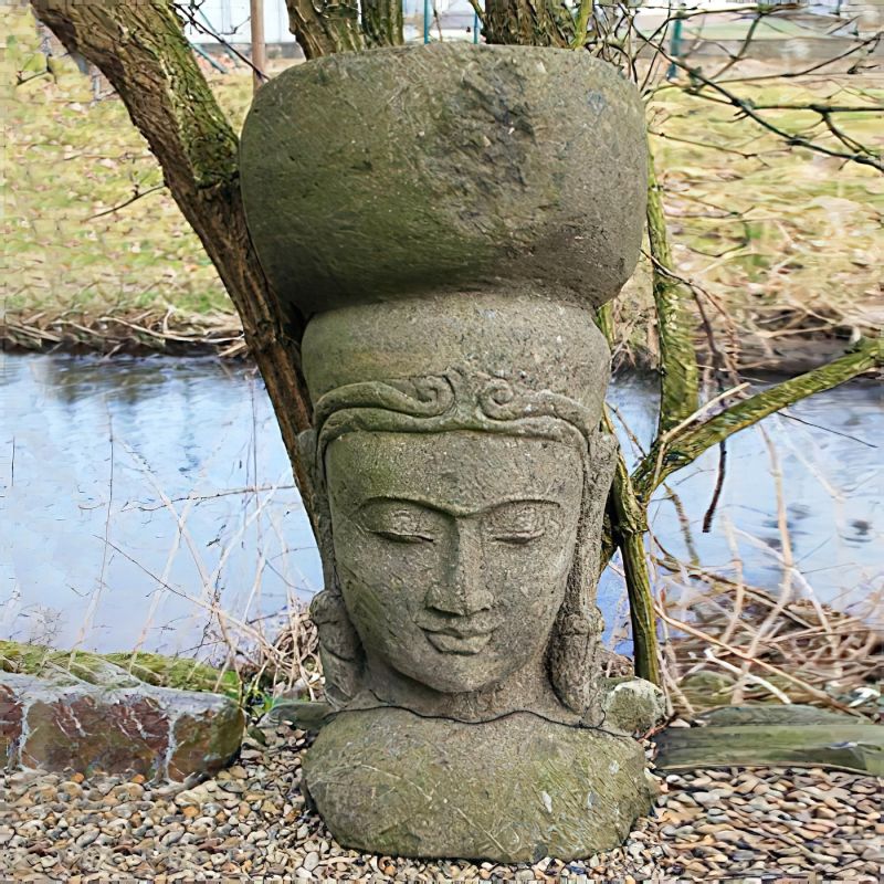 Grande statua di testa in pietra come decorazione da giardino vicino a un ruscello e un albero