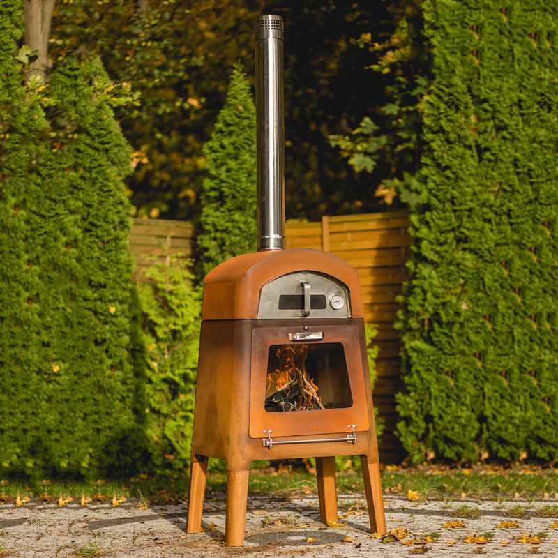 Forno da giardino in metallo con fuoco visibile, circondato da siepi verdi nel giardino