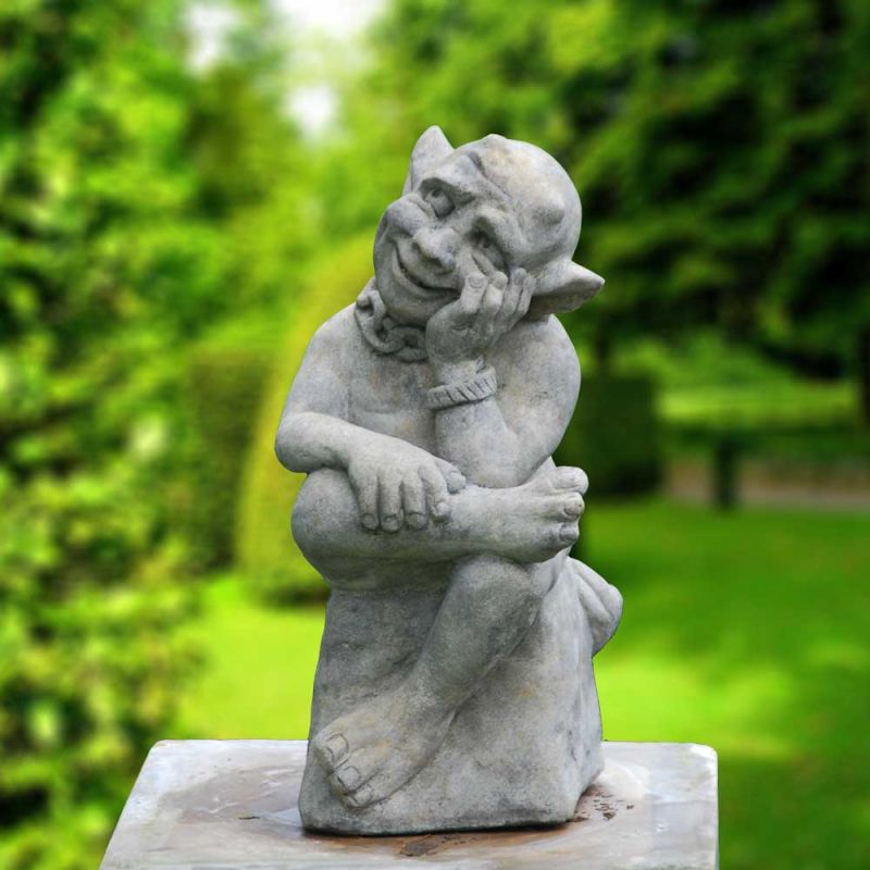 Figurine grise de gargouille assise, pensive, sur une pierre dans un jardin verdoyant