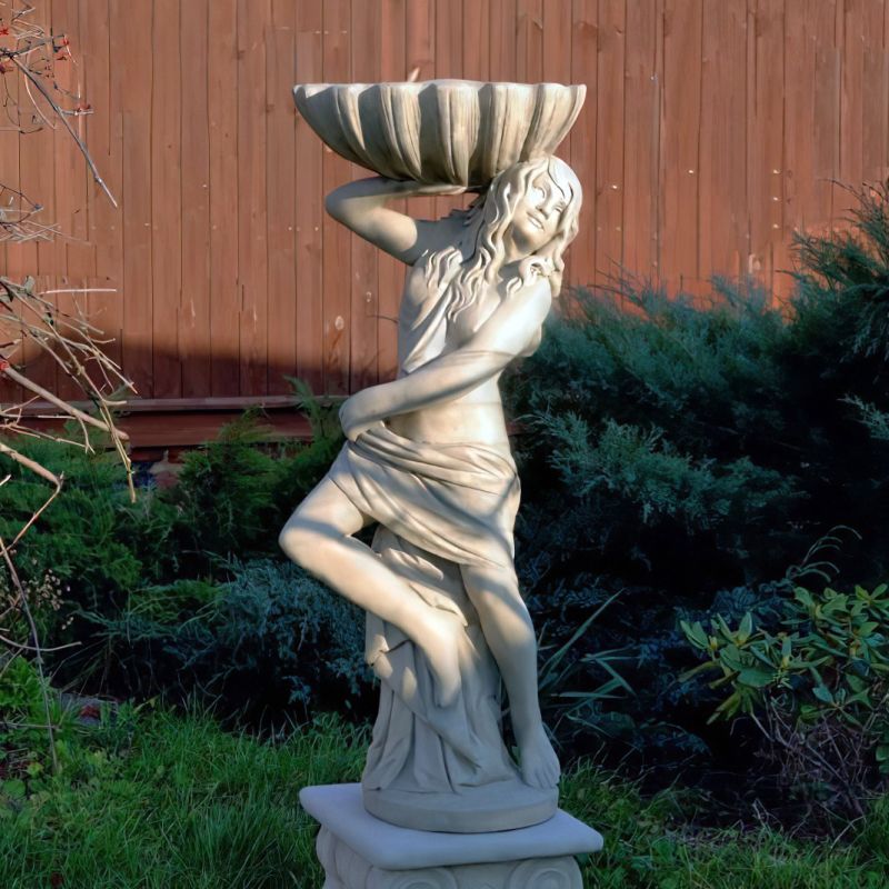 Fontaine de jardin blanche avec figure féminine devant une clôture en bois et des plantes vertes
