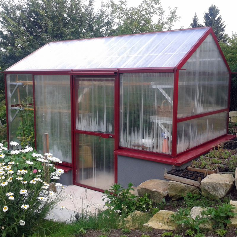 Serre avec cadre rouge en matériau transparent dans un jardin entouré de verdure et de fleurs