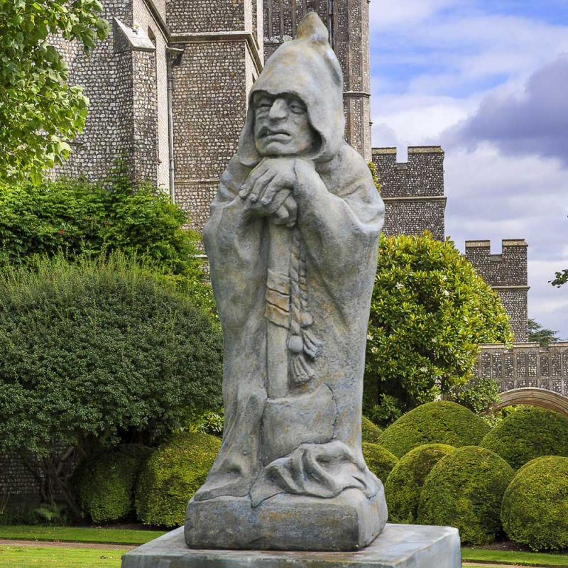 Jardin avec une statue en pierre d'un moine devant un château avec des arbustes et arbres verts